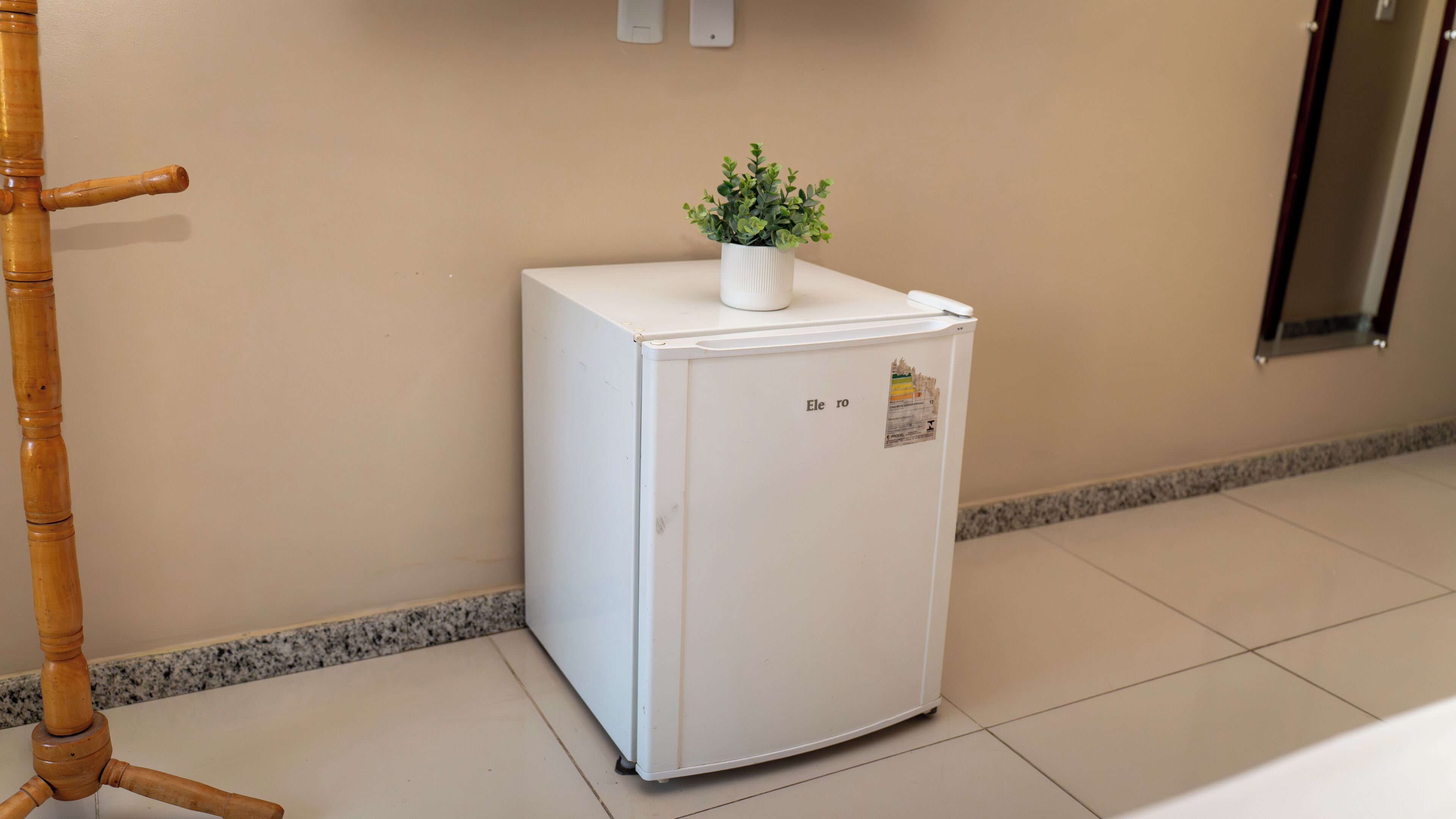 A small white refrigerator with a plant on top, located in a hallway with a tiled floor and a wooden chair nearby.