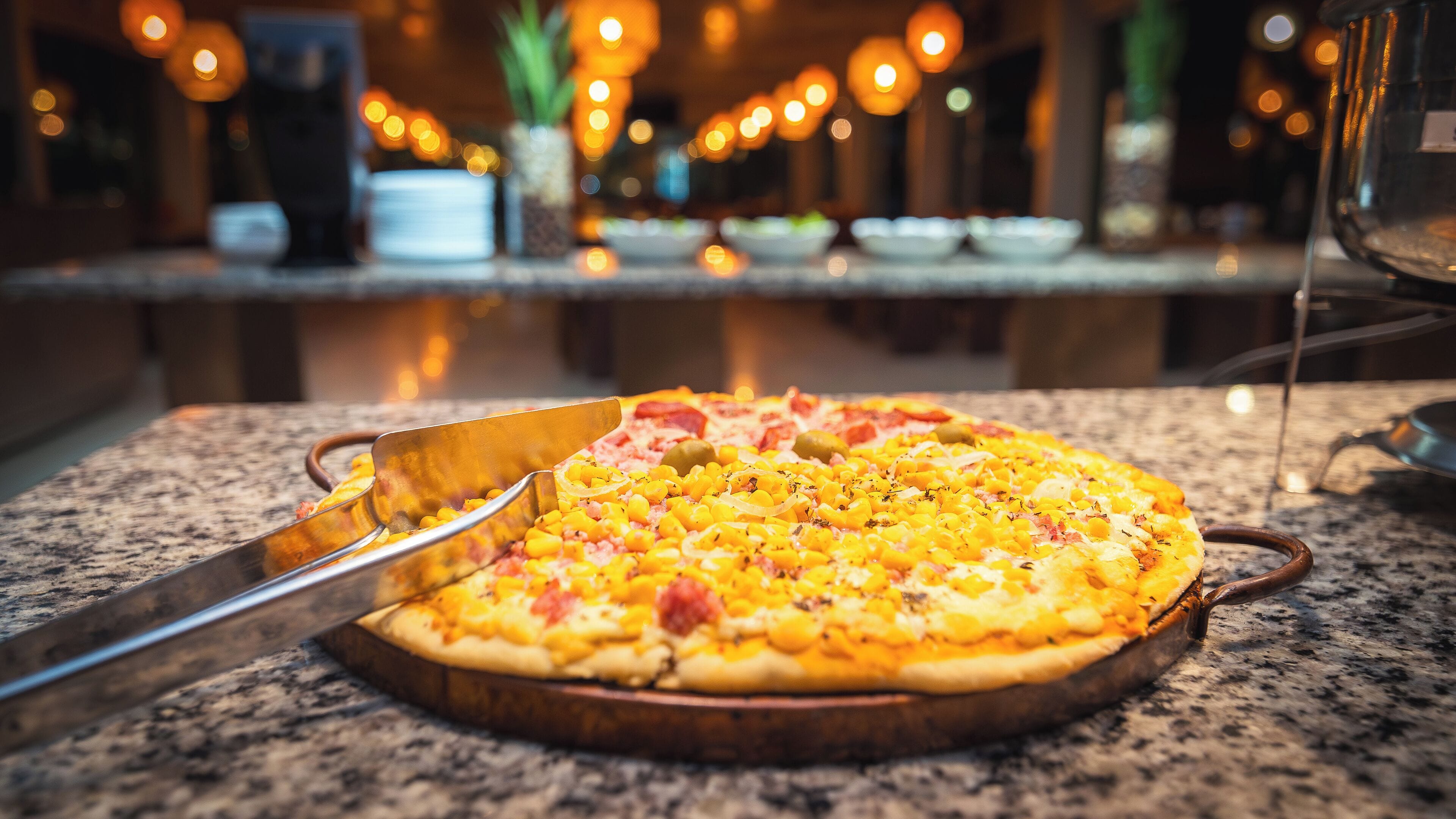 A pizza on a wooden board with a pizza cutter and a glass of water on a marble countertop.