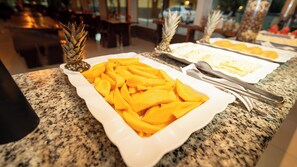 A buffet table with plates of sliced mangoes and pineapples, and a pair of tongs on the granite countertop. - Pousada Itajuipe (Itabuna)