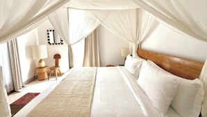 A spacious bedroom with a canopy bed, a wooden headboard, a nightstand with a lamp, and a small wooden chair. - Villa Hummingbird Ubud (Ubud)