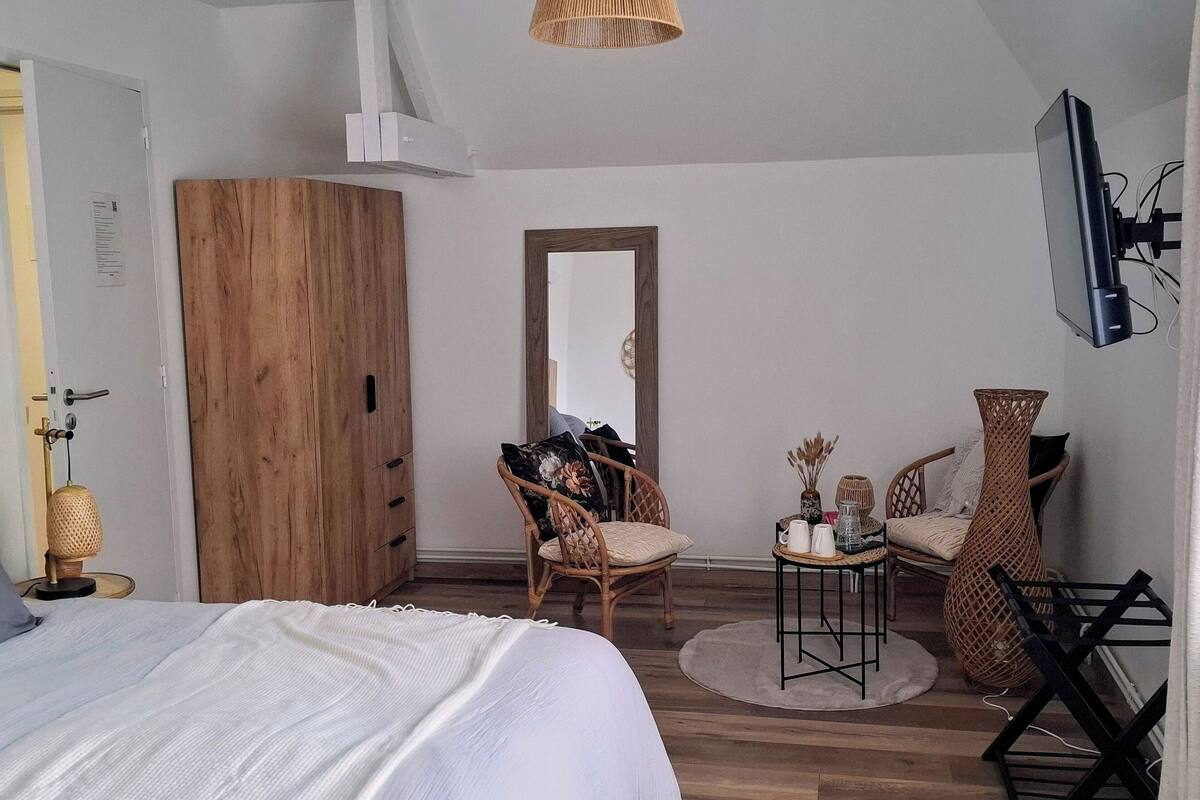 A bedroom with a bed, a wooden wardrobe, a mirror, wicker chairs, a small table with items, and a TV mounted on the wall.