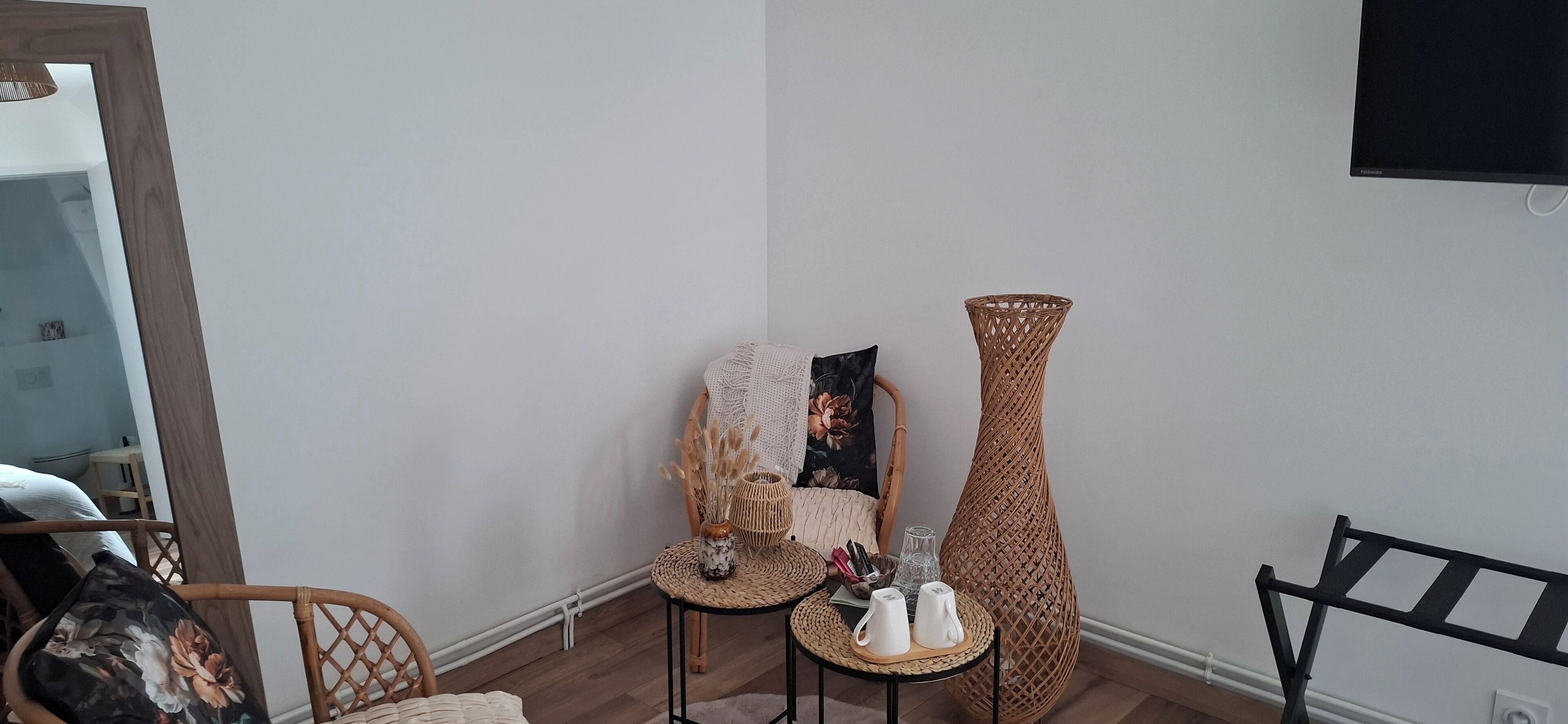A room with a wicker chair, a small table with cups, a tall vase, and a flat-screen TV mounted on the wall.