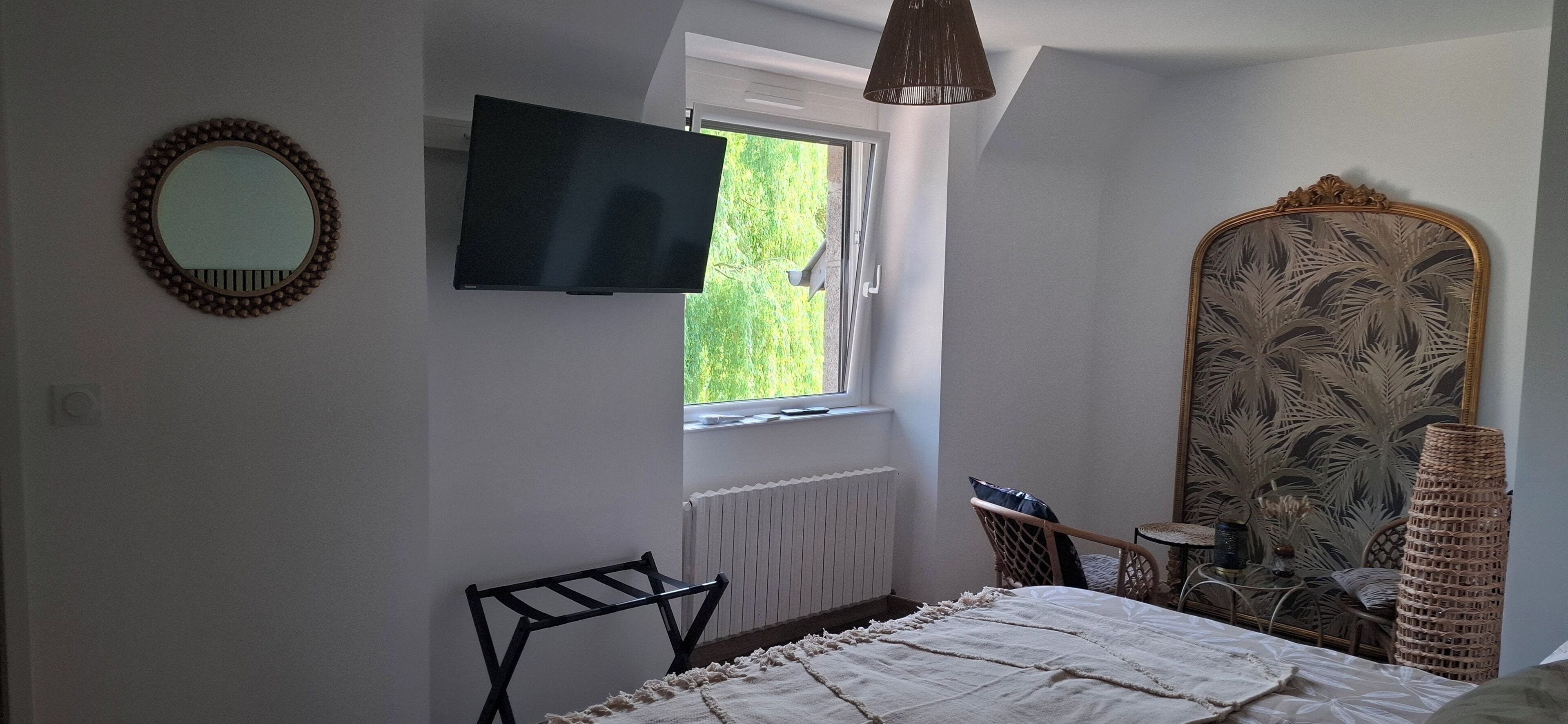 A bedroom with a bed, a TV mounted on the wall, a window, a radiator, a chair, and a decorative mirror.