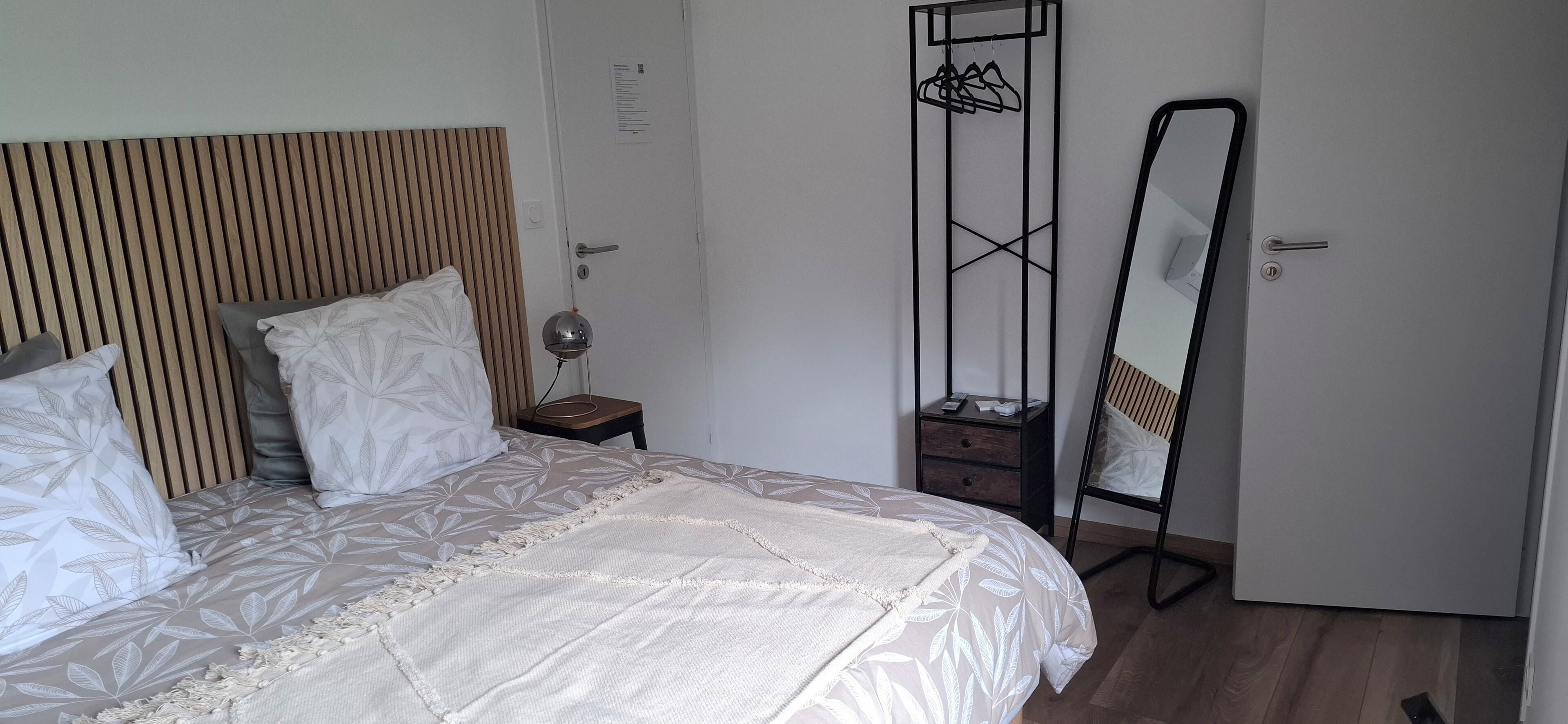 A neatly made bed with a patterned bedspread, a bedside lamp, a wooden headboard, a wardrobe with a mirror, and a small nightstand.