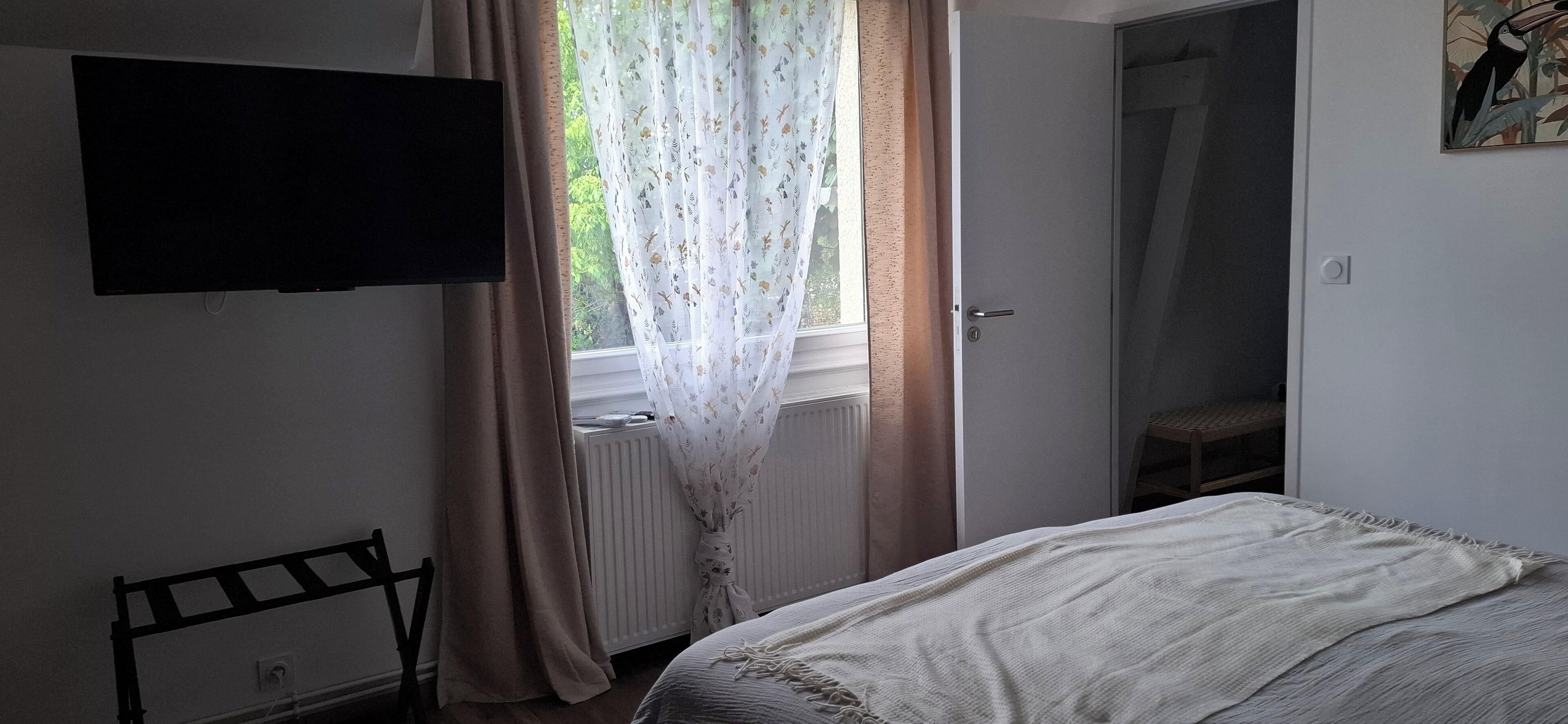Un dormitorio con cama, radiador, televisión montada en la pared y ventana con cortinas.