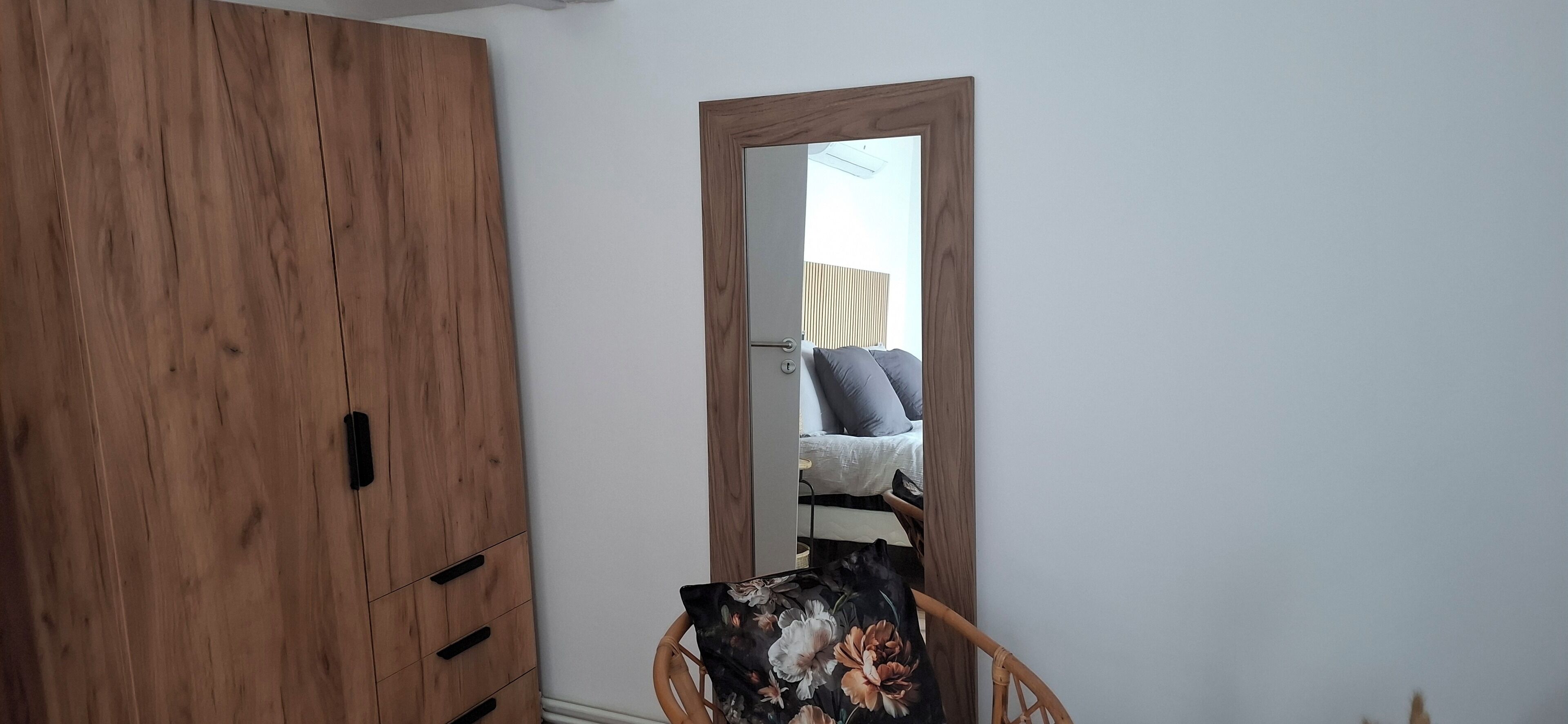 Una habitación con un armario de madera, un espejo que refleja una cama con almohadas y una silla con cojín floral.