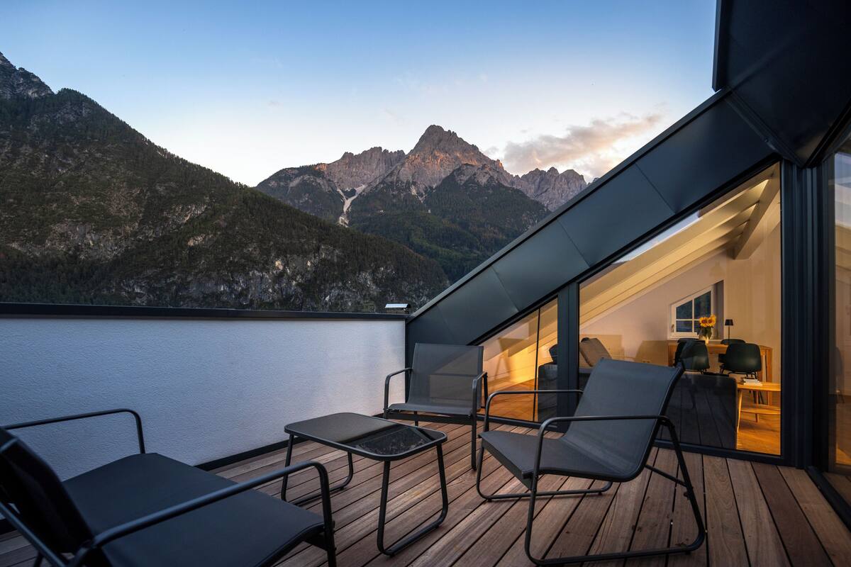 A balcony with a wooden deck, black metal chairs, and a glass door leading to an interior space with a desk and chair.