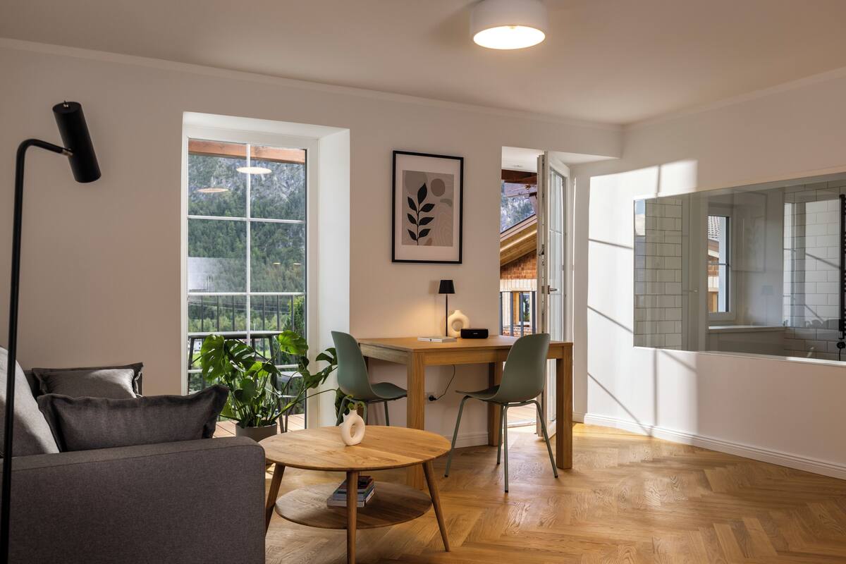 A modern living room with a sofa, a dining table with chairs, a floor lamp, and a large window with a view of greenery.