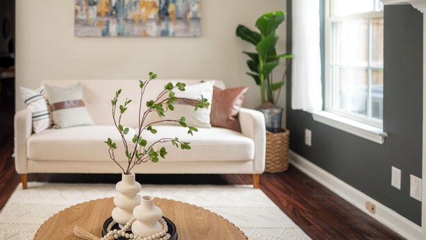 A living room with a white sofa, a coffee table with a vase and beads, a large abstract painting on the wall, and a plant in a basket.