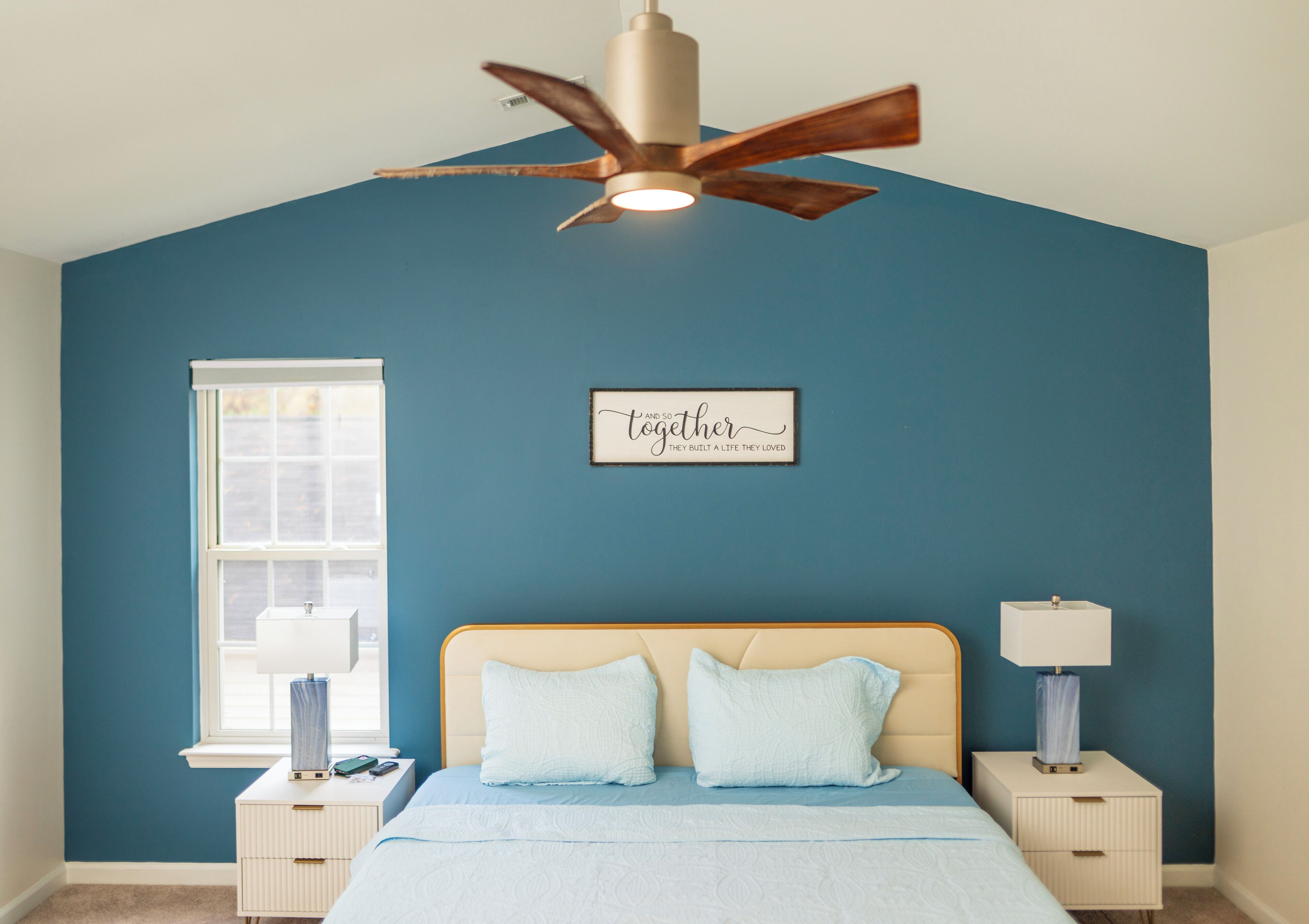 A bedroom with a bed, two bedside tables, a ceiling fan, a window, and a wall with a motivational quote.