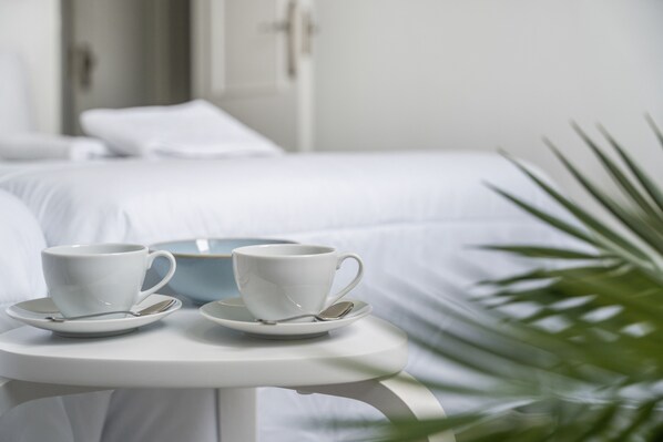 A bed with white bedding, a white table with two cups and saucers, and a green plant in the foreground.