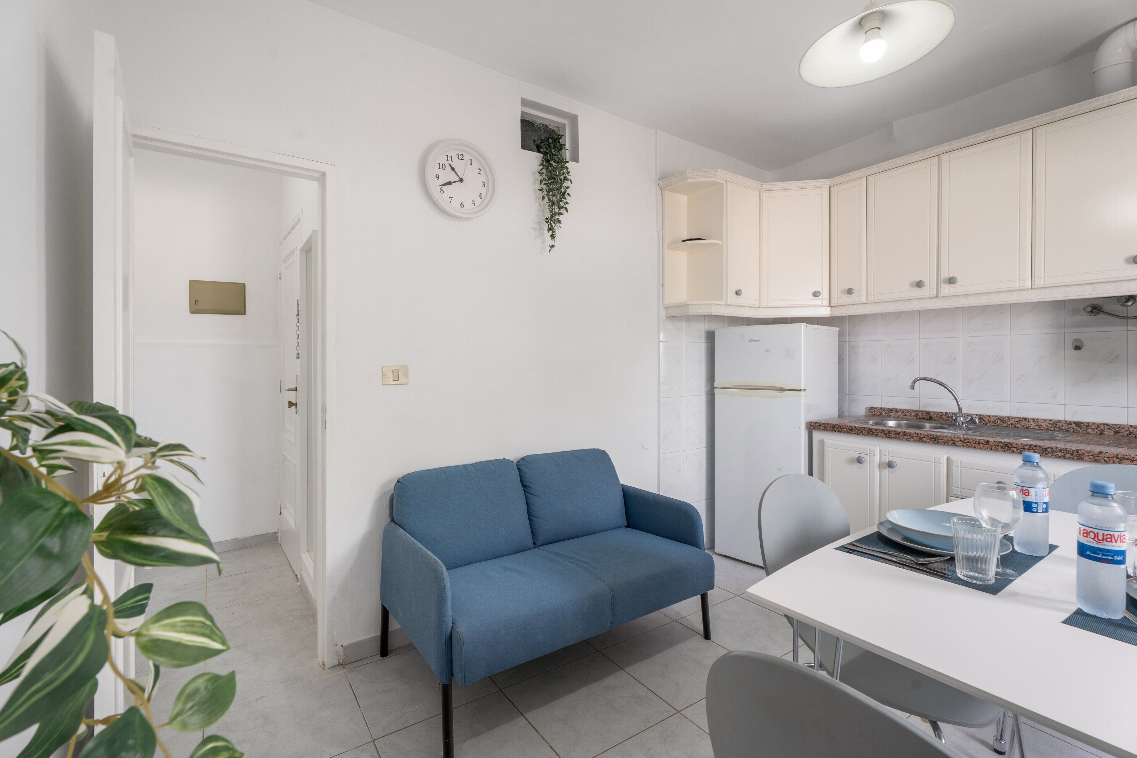 Une petite cuisine avec des meubles blancs, une horloge et une plante.