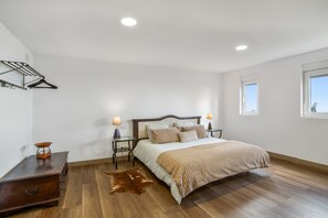 A bedroom with a wooden floor, a bed with a beige blanket, two bedside lamps, a wooden dresser, a hanging clothes rack, and a window.