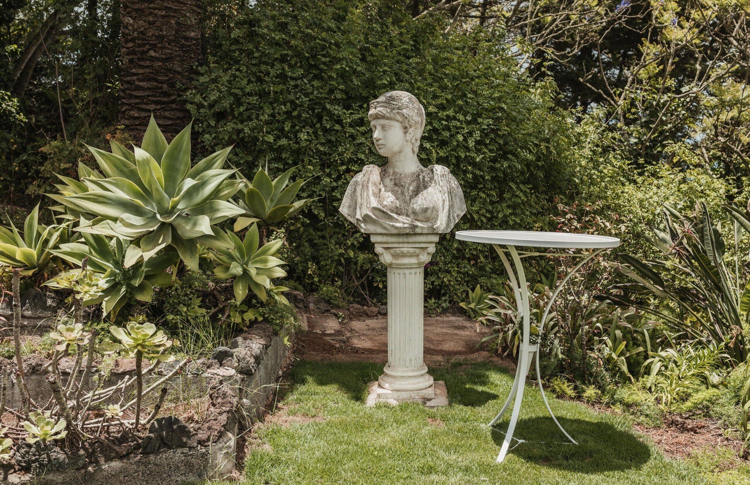 Un jardin agrémenté d’un buste sculpté, posé sur un piédestal, et entouré de différentes plantes ainsi que d’une petite table.