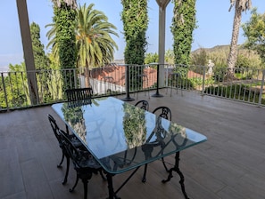 A glass-top table with chairs on a balcony with a view of greenery and a fence.