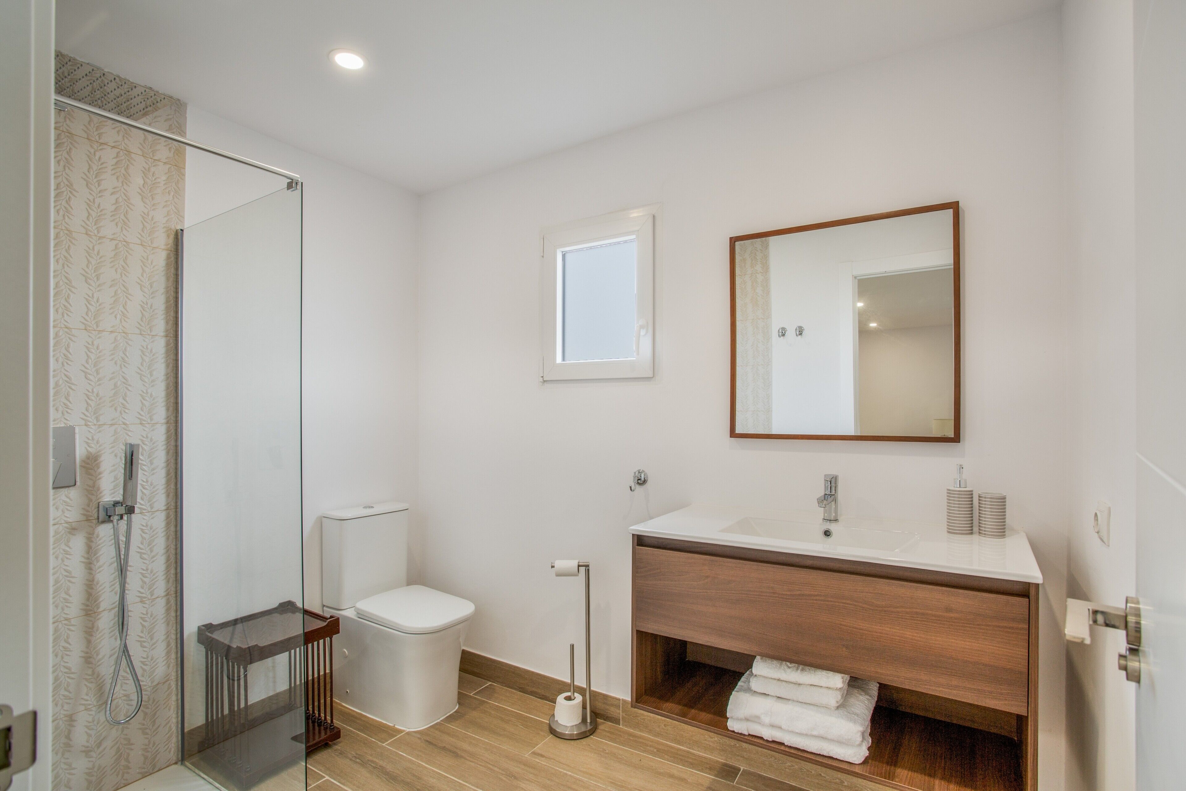 A modern bathroom with a glass shower enclosure, a toilet, a sink with a wooden vanity, and a mirror.