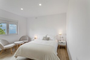Un dormitorio moderno con una cama grande, dos mesitas de noche, una silla y una ventana con vistas a árboles.