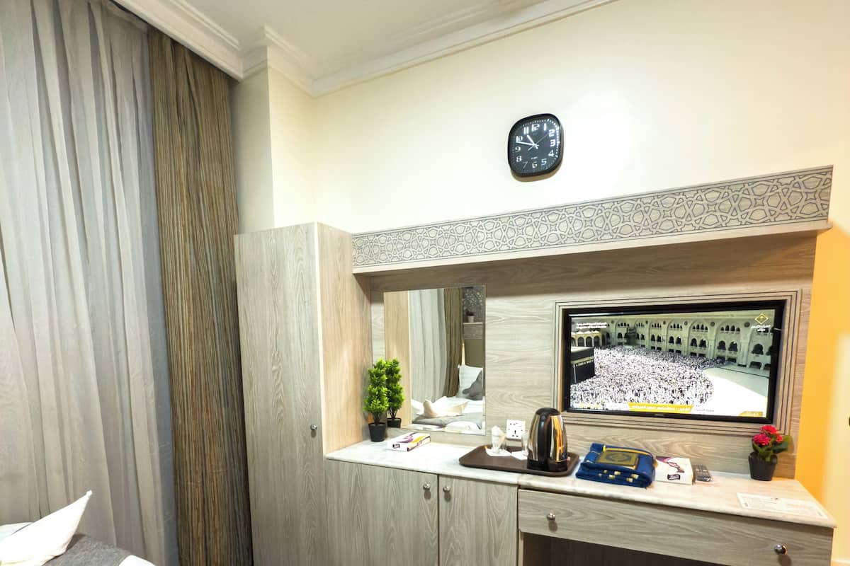 A hotel room with a bed, a desk, a mirror, a clock, and a television displaying a crowd.