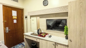 Une chambre d’hôtel de taille modeste, dotée d’une porte en bois, d’un petit bureau avec un écran d’ordinateur, une horloge et une petite plante.