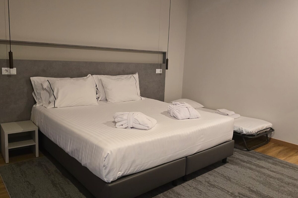 A modern hotel room with a large bed, a bedside table, a small stool, and a grey carpet.