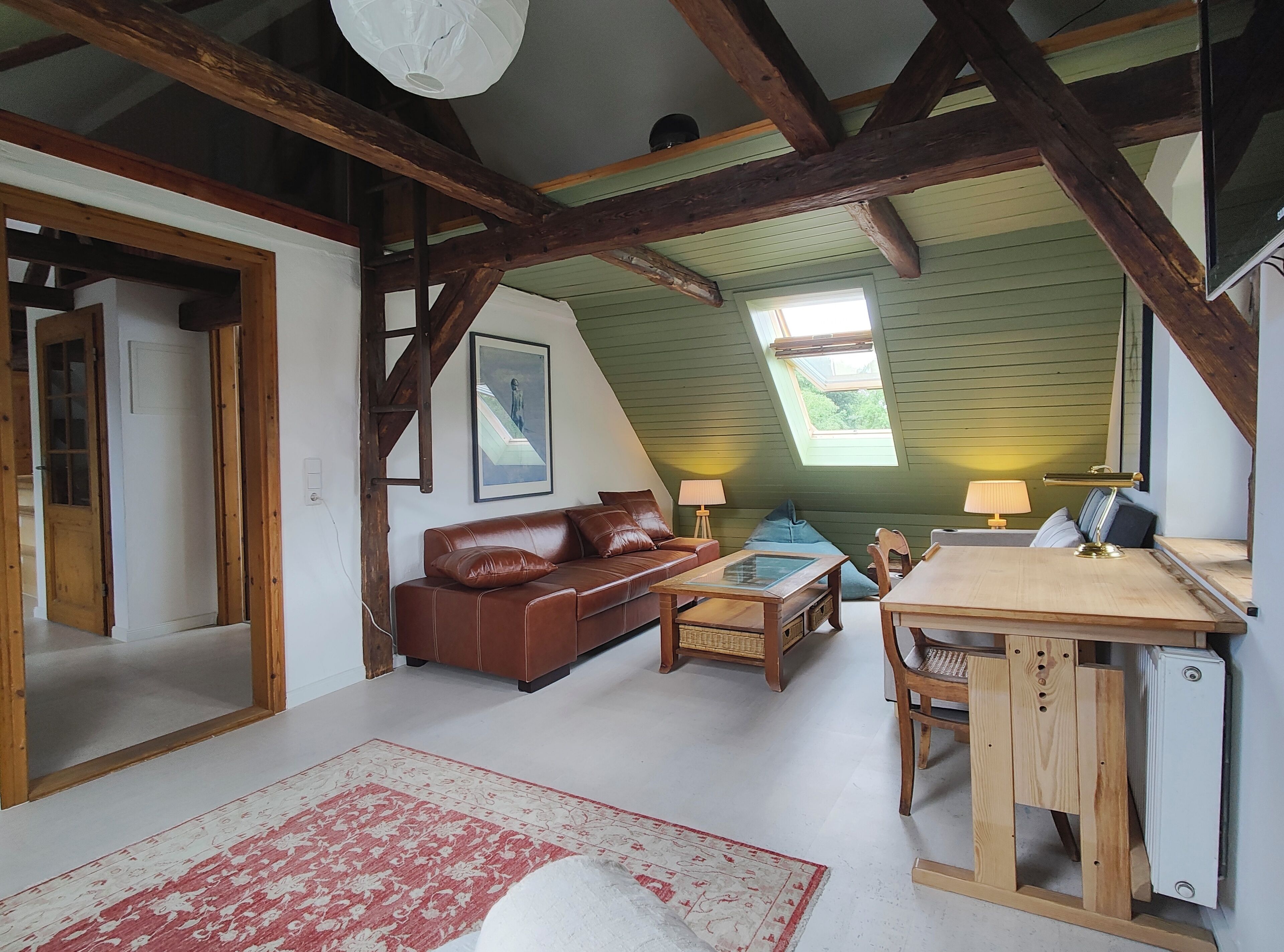 Ein gemütliches Dachzimmer mit schrägem Dach, Holzbalken, einem braunen Ledersofa, einem kleinen Tisch mit Lampe und Blick durch ein Dachfenster.