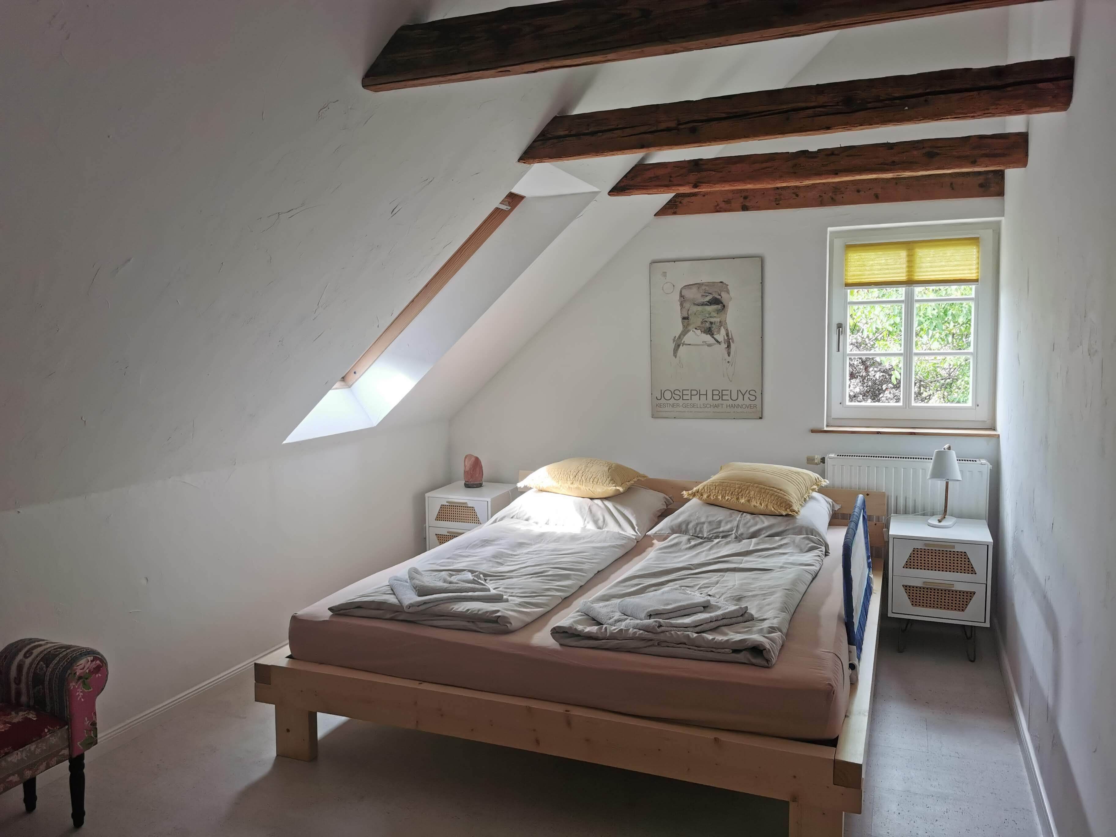 A bedroom with a sloped ceiling, a bed with two pillows, a nightstand, a chair, and a window with blinds.