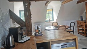Una cuina acollidora amb taula de fusta, cafetera i un aparell Siemens. Hi ha dues finestres amb cortines, una cadira de fusta i un petit armari.