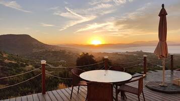 Una terraza de madera con una mesa redonda y sillas, con vista a un paisaje pintoresco al atardecer.