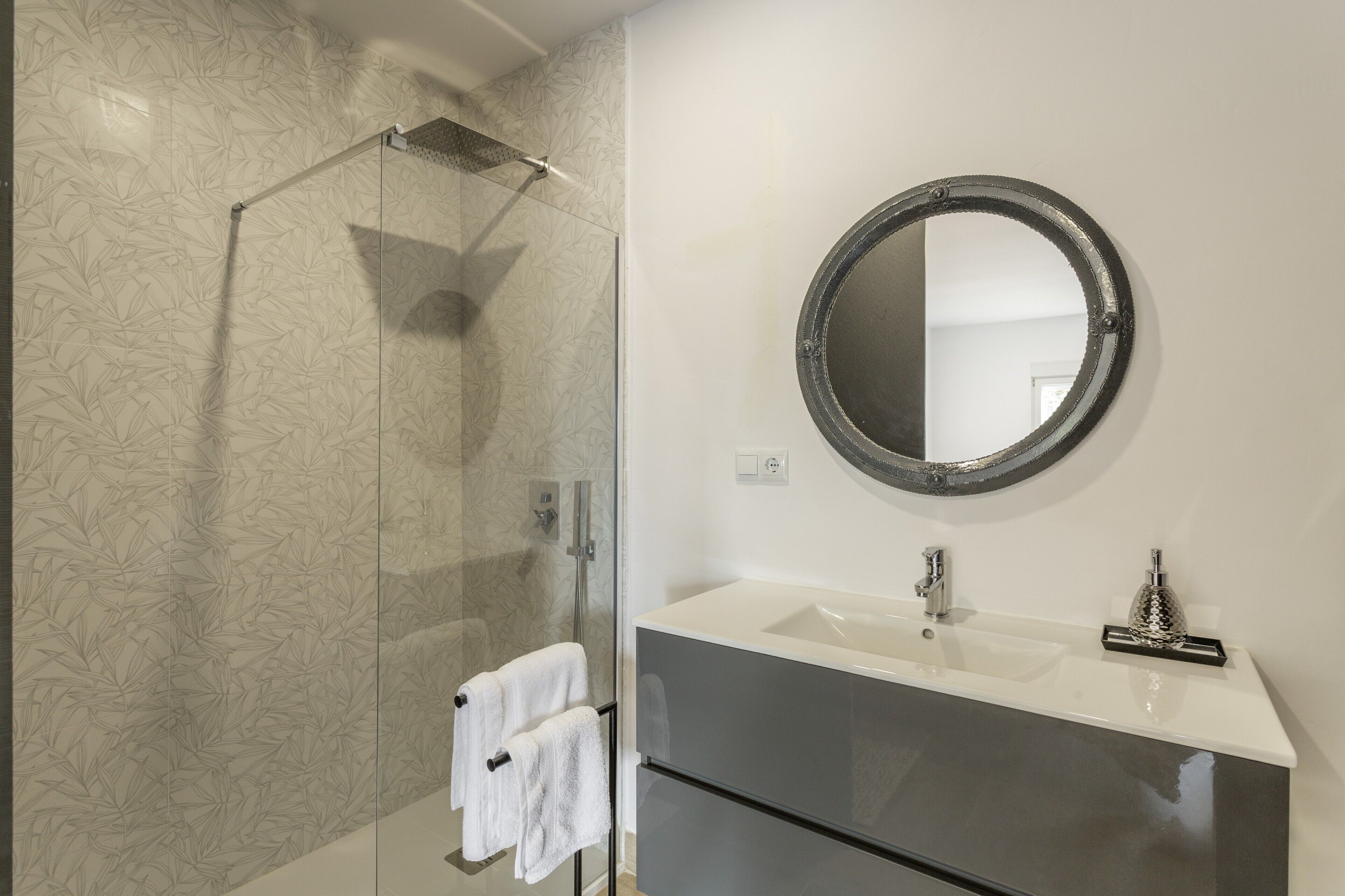 A bathroom with a glass shower enclosure, a white sink with a round mirror, and a towel rack with two towels.