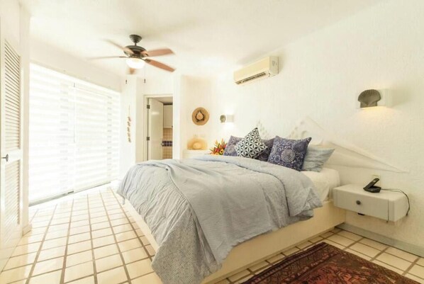 A bedroom with a bed, a nightstand, a ceiling fan, a wall-mounted air conditioner, and a window with blinds.