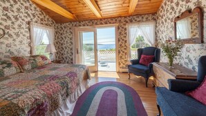 Un dormitorio con techo de madera, papel tapiz floral, una cama con colcha colorida, un sillón azul y vistas al exterior a través de un ventanal.