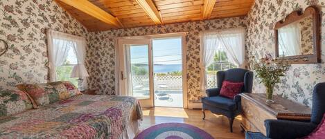 A bedroom with a wooden ceiling, floral wallpaper, a bed with a colorful quilt, a blue armchair, and a view of the outdoors through a glass door.
