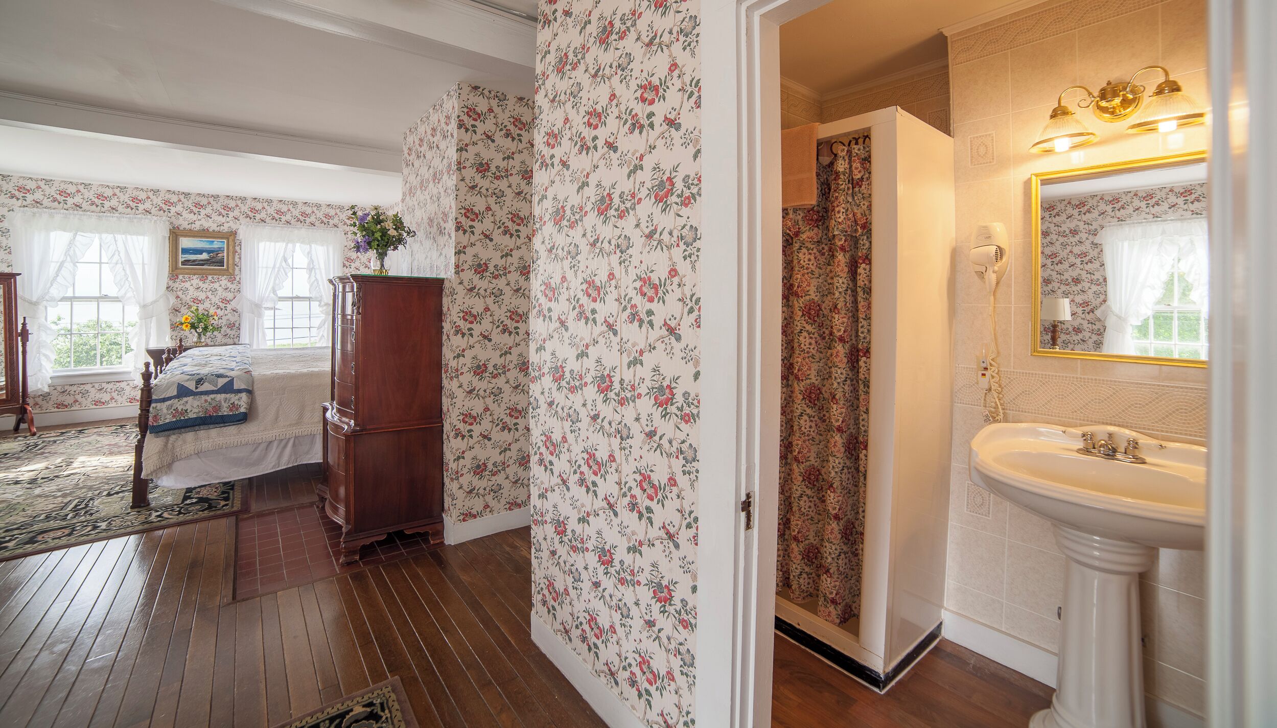 A bedroom with floral wallpaper, a wooden dresser, a bed with a quilt, and a bathroom with a sink and mirror.