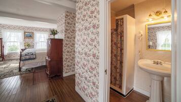 A bedroom with floral wallpaper, a wooden dresser, a bed with a quilt, and a bathroom with a sink and mirror.