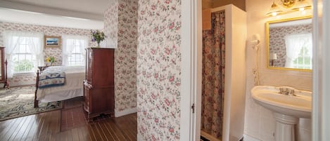 A bedroom with floral wallpaper, a wooden dresser, a bed with a quilt, and a bathroom with a sink and mirror.