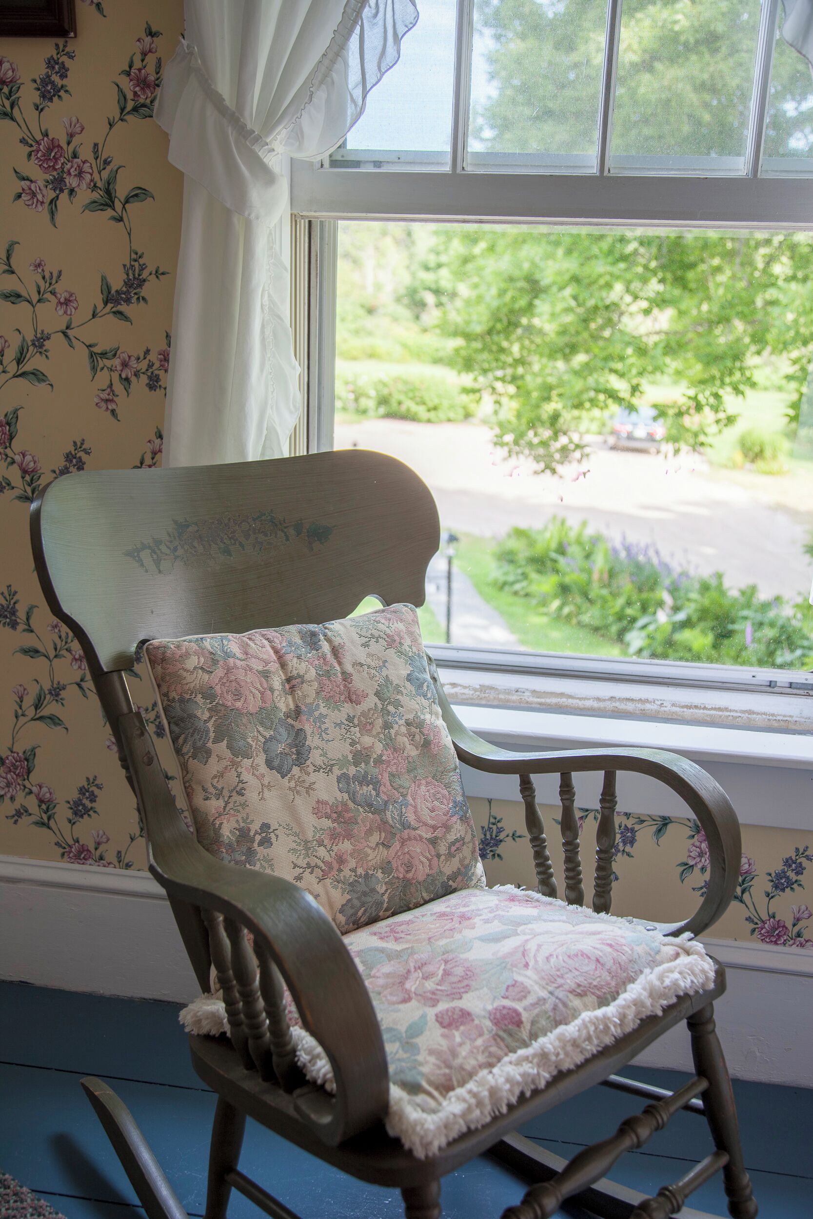 A wooden rocking chair with a floral cushion in front of a window with a view of greenery and a road.