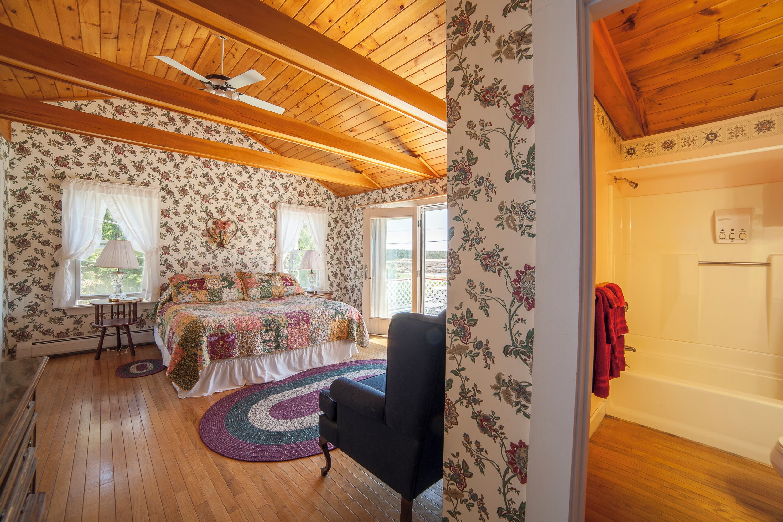 Una camera da letto con soffitto in legno, carta da parati floreale, un letto con copripiumino colorato, una sedia nera e vista sull'esterno.