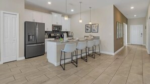 A modern kitchen with white cabinets, a stainless steel refrigerator, a microwave, and a bar area with four bar stools.