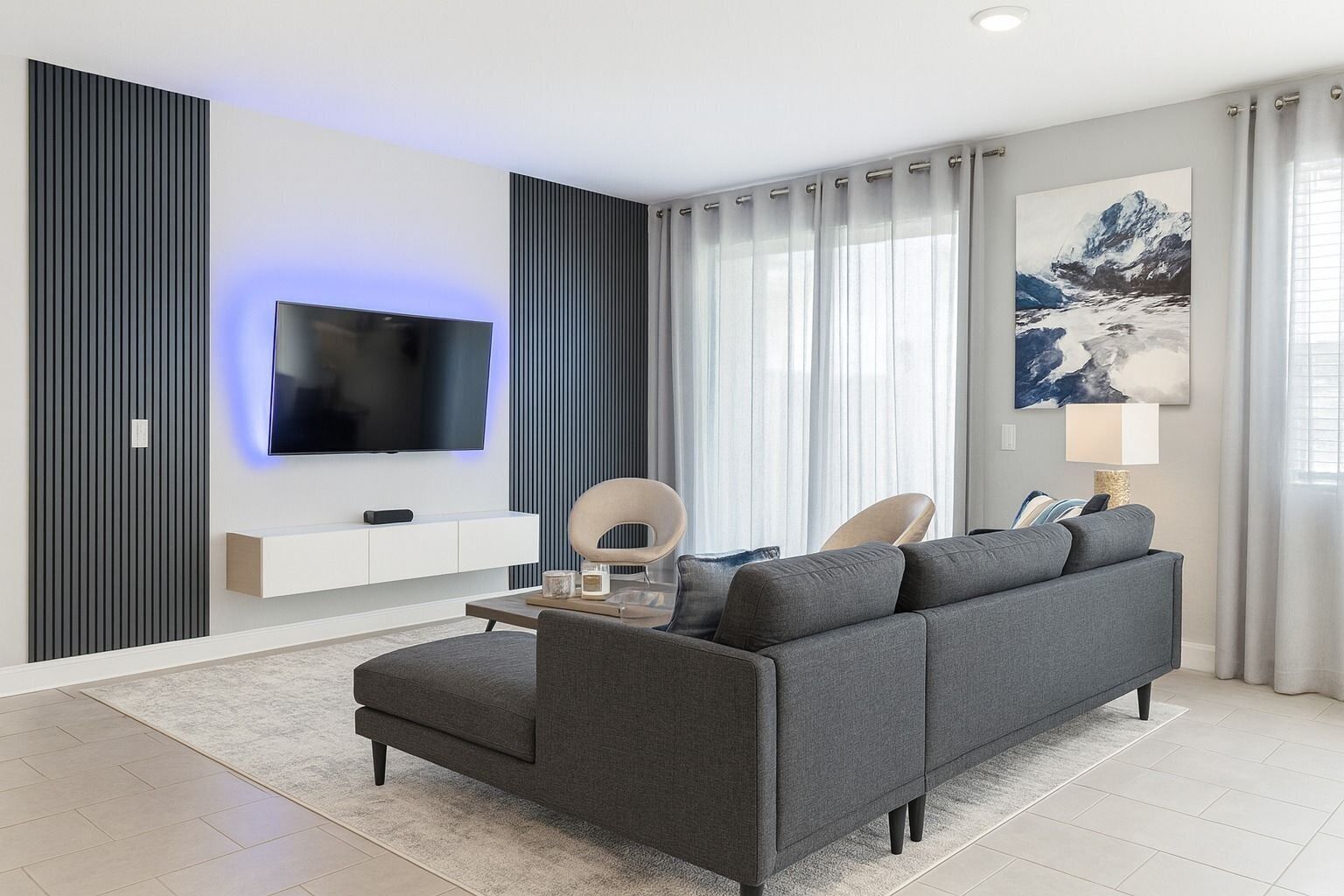 A modern living room with a grey sofa, a flat-screen TV mounted on a white wall, a decorative mirror, and a painting of a mountain landscape.