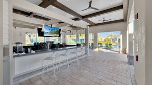 A spacious outdoor bar area with a modern design, featuring a counter with bar stools, ceiling fans, and a large TV screen displaying a scenic view.