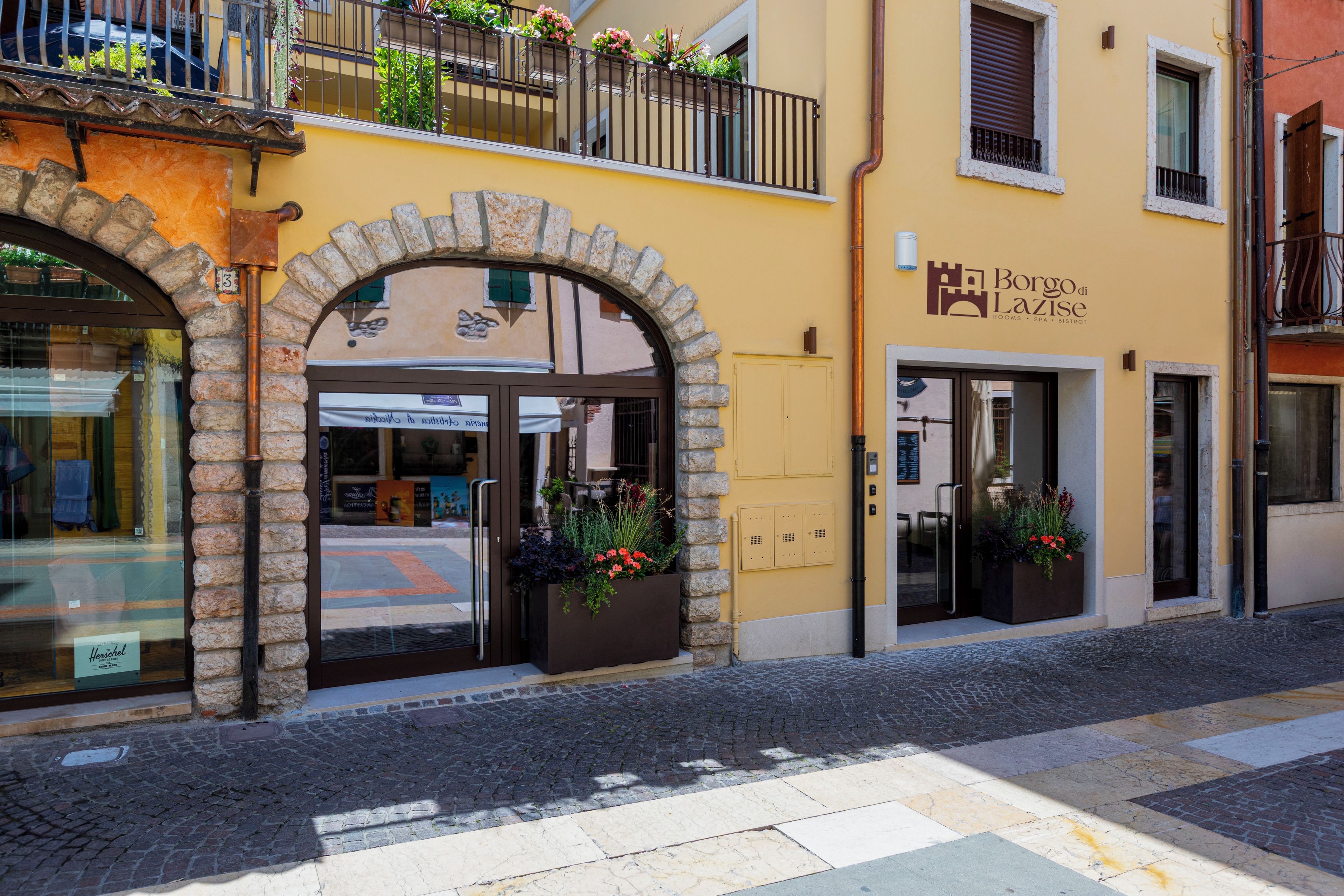 L’entrée d’un hôtel, avec une porte en arc de cercle, une façade en pierre et une enseigne indiquant « Borgo di Lazise », signalant la présence de chambres, d’un spa et d’un bistrot.