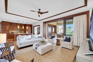 A spacious living area with a sofa, armchairs, a coffee table, and a TV. There is a kitchen in the background with wooden cabinets and pendant lighting.
