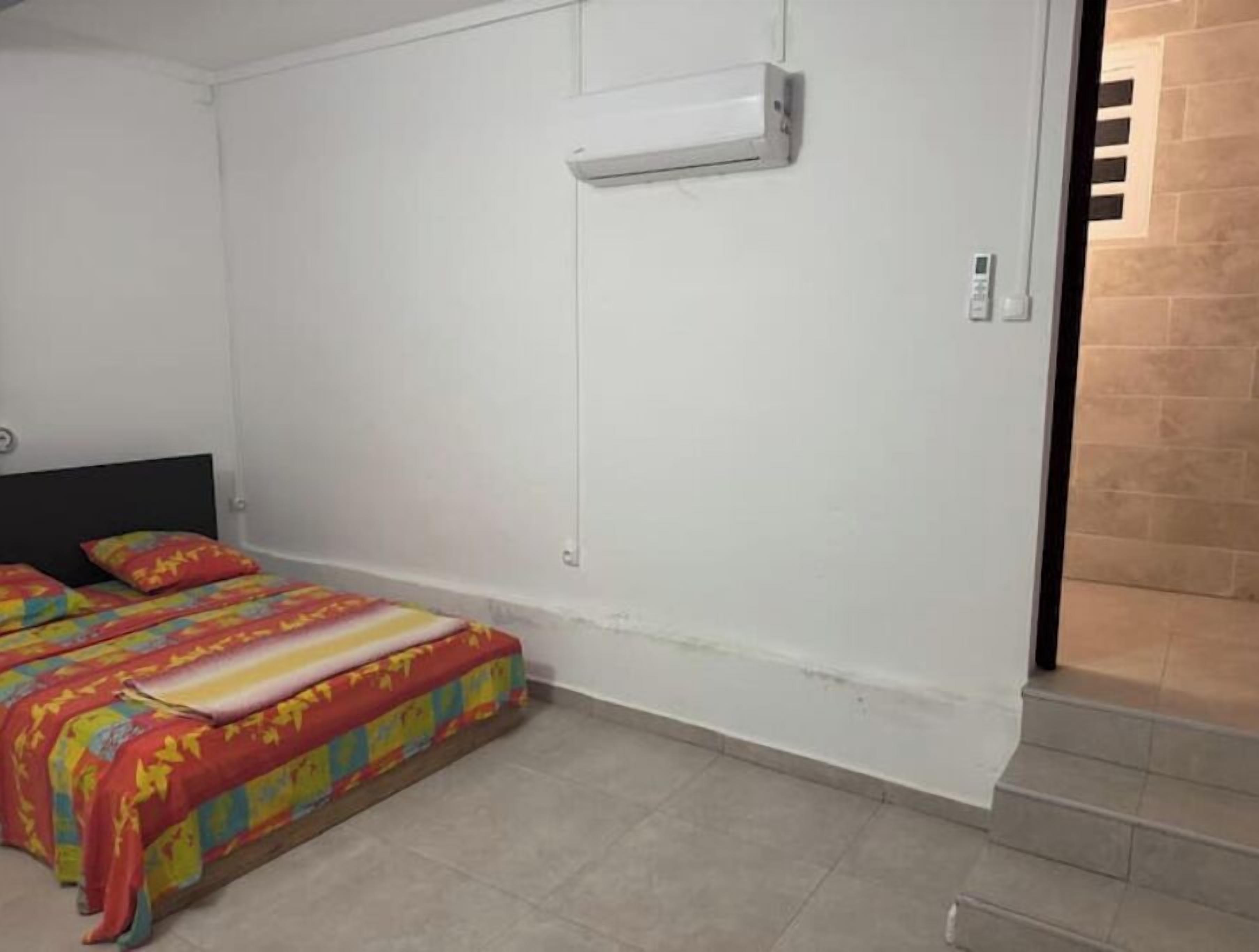 Ein Schlafzimmer mit einem Bett, das mit farbenfroher Bettwäsche bezogen ist, einer weißen Wand und einer an der Wand montierten Klimaanlage.