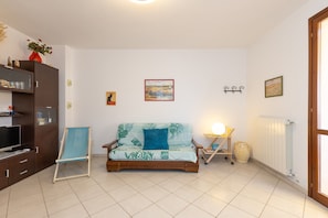 Una sala de estar con un sofá, un televisor, un mueble de madera, una mesita con una lámpara y un radiador.