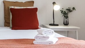 Un lit bien fait, avec un oreiller rouge et une couverture blanche, une lampe de chevet, une petite plante en pot et une pile de serviettes pliées.