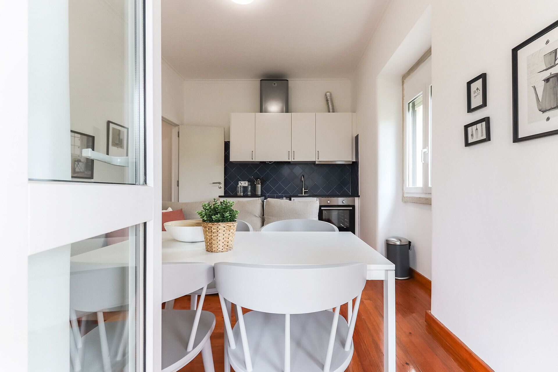 A modern kitchen with white cabinets, a black backsplash, and stainless steel appliances. There is a dining area with a white table and chairs, and a potted plant on the table.