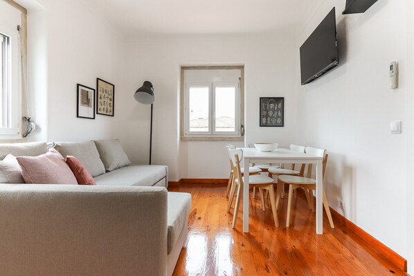 A compact living space with a sofa, a dining table with chairs, a TV mounted on the wall, and a window with a view. - Belem Boutique Viii by Homing (Lisbon)