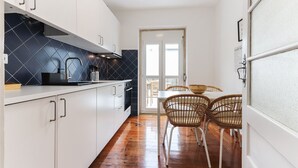 A narrow kitchen with white cabinets, a black backsplash, and a wooden floor. There is a dining area with wicker chairs and a table. - Belem Boutique IX by Homing (Lisbon)