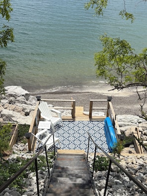 Un escalier mène à une zone de plage agrémentée d’une terrasse en bois, d’un revêtement de sol à motifs blancs et bleus, et d’un fauteuil de plage bleu.
