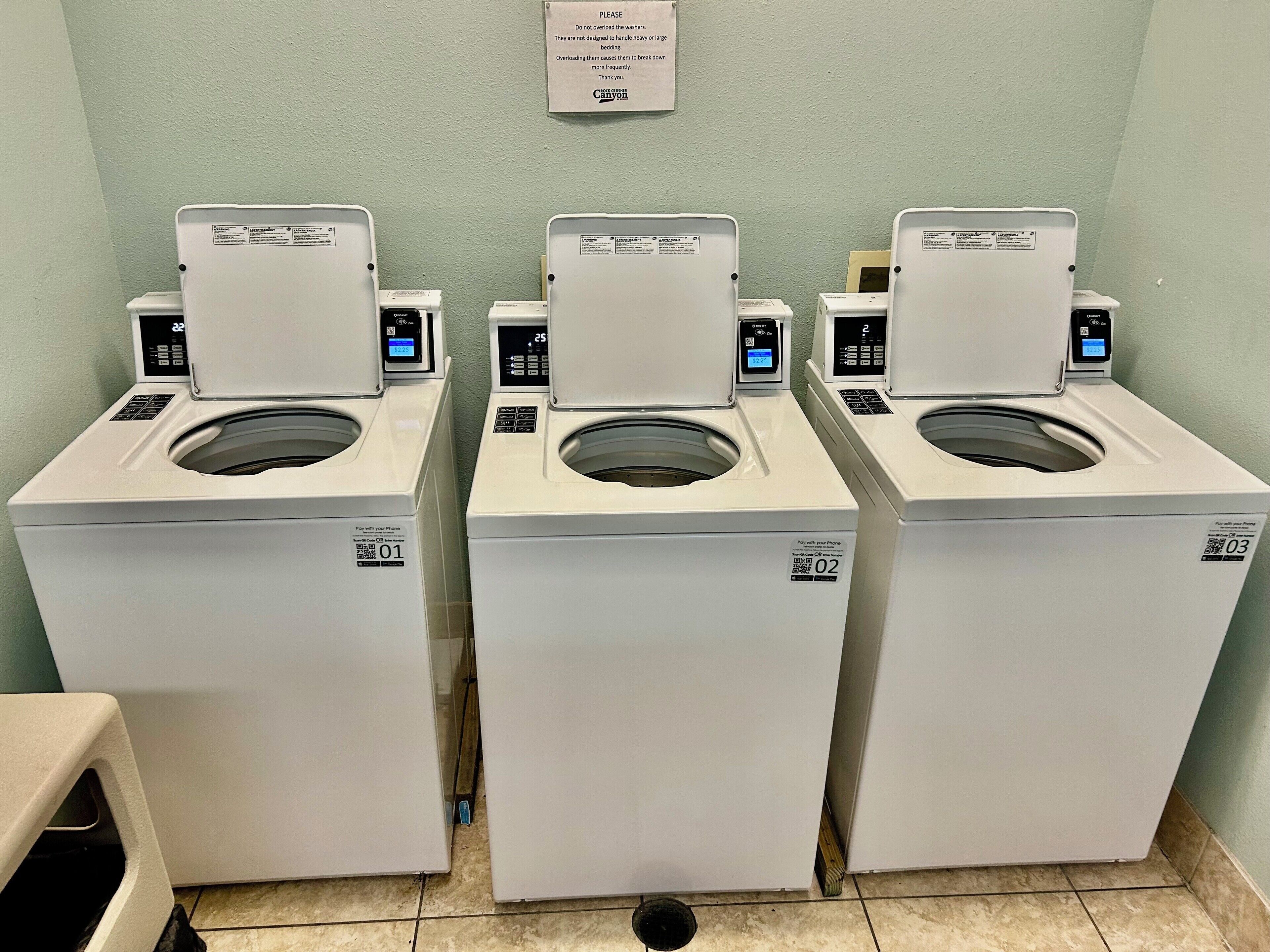 Une rangée de trois machines à laver blanches à chargement frontal se trouve dans une buanderie, et un panneau est accroché au mur.